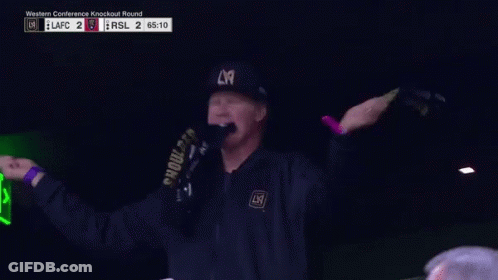 A Man Wearing A La Hat And A Scarf Holds His Fist Up GIF