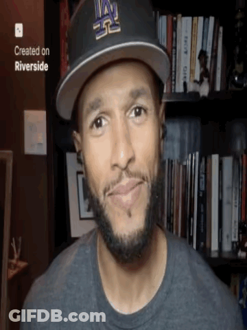 A Man Wearing A La Hat Is Smiling In Front Of A Bookshelf GIF