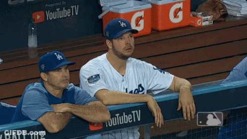 A Man Wearing A La Hat Sits In A Dugout Next To Another Man GIF