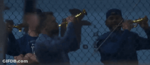 A Man Wearing A La Hat Stands Behind A Chain Link Fence While Another Man Plays A Violin GIF