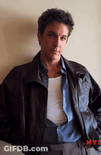 A Man Wearing A Leather Jacket And A Blue Shirt Is Standing In Front Of A Nyl Logo GIF