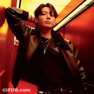 A Man Wearing A Leather Jacket And A Necklace Is Leaning Against A Red Wall . GIF