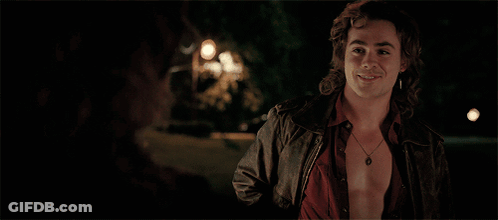 A Man Wearing A Leather Jacket And A Necklace Smiles At Another Man GIF