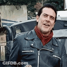 A Man Wearing A Leather Jacket And A Red Scarf Is Standing In Front Of A Car And Smiling . GIF
