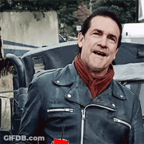 A Man Wearing A Leather Jacket And A Red Scarf Is Standing In Front Of A Vehicle . GIF