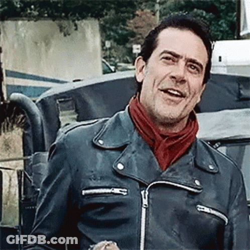 A Man Wearing A Leather Jacket And A Scarf Is Standing In Front Of A Car . GIF