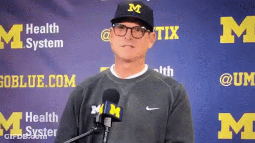 A Man Wearing A M Hat And Glasses Is Standing In Front Of A M Banner . GIF