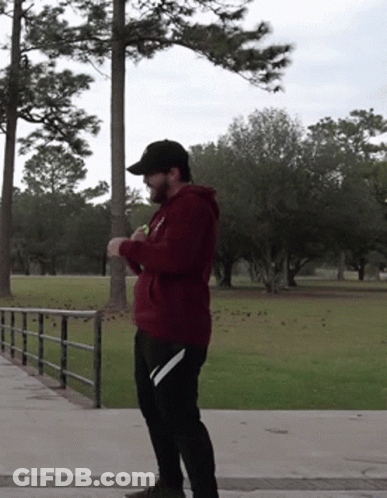A Man Wearing A Maroon Hoodie And Black Hat Stands In A Park GIF
