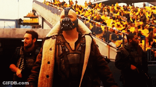 A Man Wearing A Mask And A Fur Coat Stands In Front Of A Crowd Of People GIF