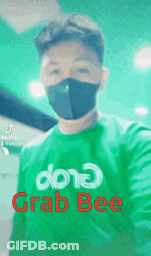 A Man Wearing A Mask And A Green Shirt That Says Grab Bee Is Taking A Selfie . GIF