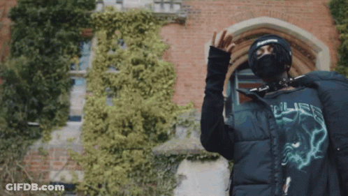 A Man Wearing A Mask And A Supreme Hat Stands In Front Of A Building GIF
