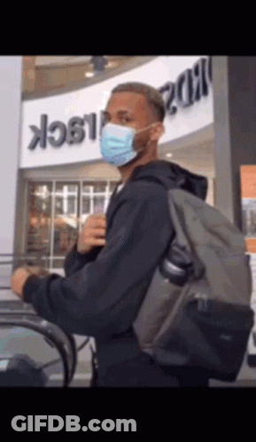 A Man Wearing A Mask And Carrying A Backpack Is Riding An Escalator GIF