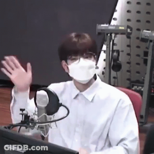 A Man Wearing A Mask And Glasses Is Sitting In Front Of A Microphone And Waving . GIF