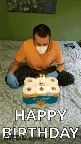 A Man Wearing A Mask And Gloves Is Sitting On A Bed With A Birthday Cake Made Out Of Toilet Paper Rolls . GIF