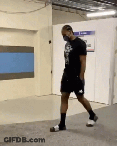 A Man Wearing A Mask And Shorts Is Walking In A Room . GIF