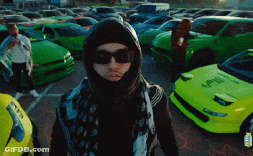 A Man Wearing A Mask And Sunglasses Is Standing In Front Of A Parking Lot Full Of Green Cars GIF