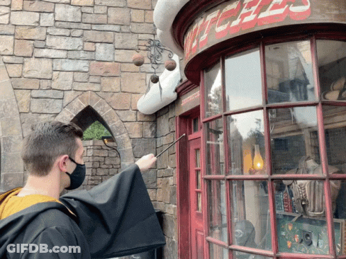 A Man Wearing A Mask Holds A Wand In Front Of A Witches Shop GIF