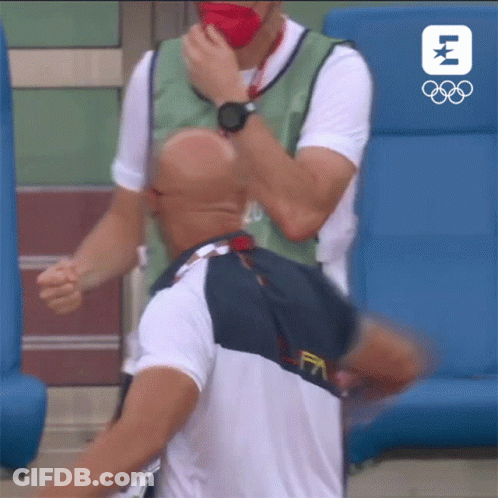 A Man Wearing A Mask Is Being Helped By Another Man With The Olympic Logo In The Background GIF