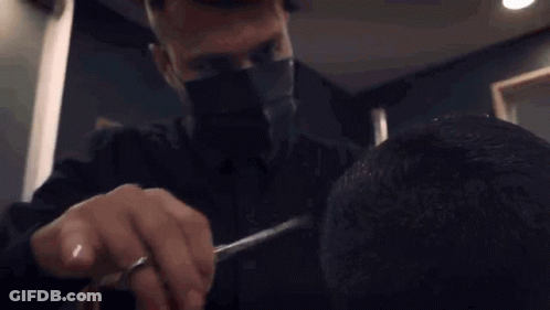 A Man Wearing A Mask Is Cutting A Man 'S Hair With A Pair Of Scissors . GIF
