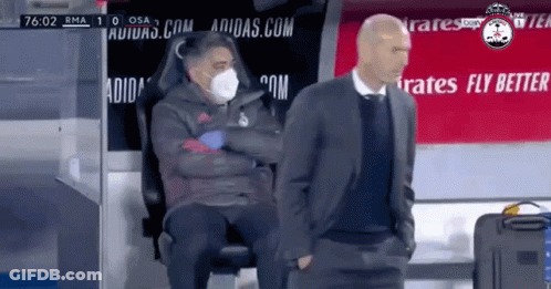 A Man Wearing A Mask Is Sitting Next To Another Man In A Suit GIF