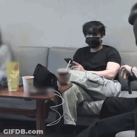 A Man Wearing A Mask Is Sitting On A Couch Looking At His Cell Phone GIF