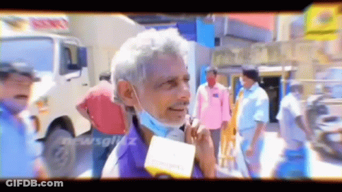 A Man Wearing A Mask Is Talking Into A Microphone While Standing In A Crowd . GIF