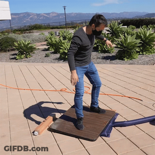 A Man Wearing A Mask Stands On A Wooden Table Outside GIF