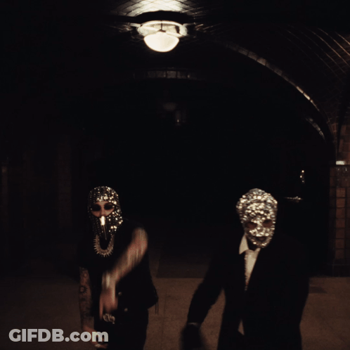 A Man Wearing A Mask With Rhinestones On It Stands Next To Another Man In A Suit GIF