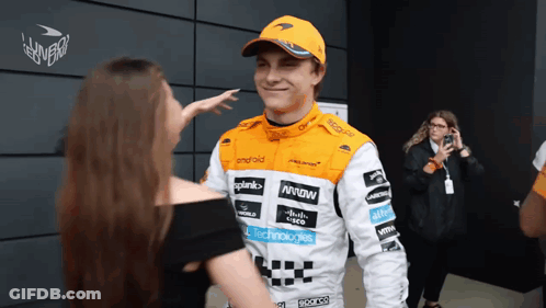 A Man Wearing A Mclaren Racing Suit Talks To A Woman GIF
