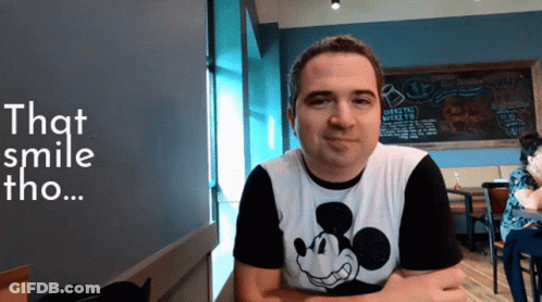A Man Wearing A Mickey Mouse Shirt Is Smiling In Front Of A Chalkboard That Says That Smile Tho GIF
