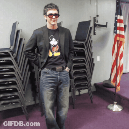 A Man Wearing A Mickey Mouse Shirt Stands In Front Of Stacks Of Chairs GIF