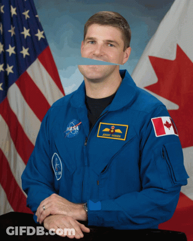 A Man Wearing A Nasa Uniform Stands In Front Of A Canadian And American Flag GIF