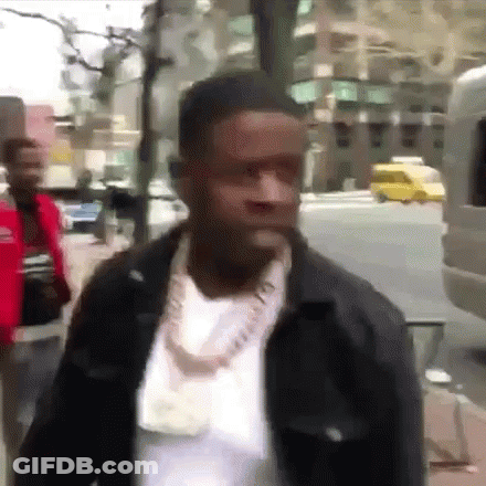 A Man Wearing A Necklace And A Jacket Is Walking Down A Street . GIF
