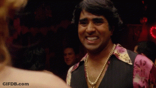 A Man Wearing A Necklace And A Pink Shirt Is Smiling At A Woman In A Club . GIF