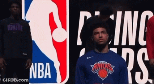 A Man Wearing A New York Knicks Shirt Is Standing In Front Of A Nba Logo GIF