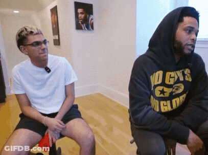 A Man Wearing A No Cys Hoodie Sits Next To A Man Wearing Shorts GIF