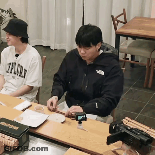 A Man Wearing A North Face Jacket Sits At A Table With Another Man GIF