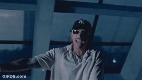 A Man Wearing A Ny Hat And Sunglasses Stands In Front Of A Window GIF