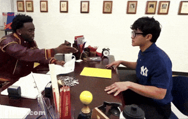 A Man Wearing A Ny Yankees Shirt Talks To Another Man At A Desk GIF