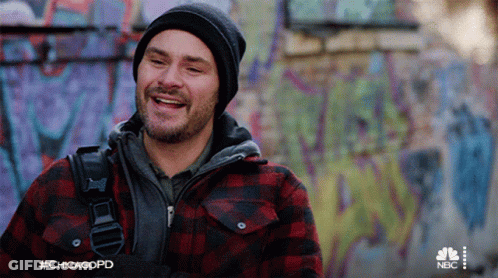A Man Wearing A Plaid Jacket And A Beanie Is Smiling In Front Of A Graffiti Covered Wall .. GIF
