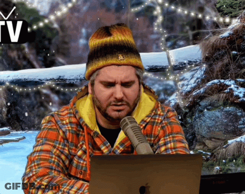 A Man Wearing A Plaid Shirt And A Beanie Is Sitting In Front Of An Apple Laptop GIF