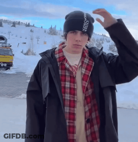 A Man Wearing A Plaid Shirt And A Black Jacket Stands In Front Of A Snowy Mountain GIF