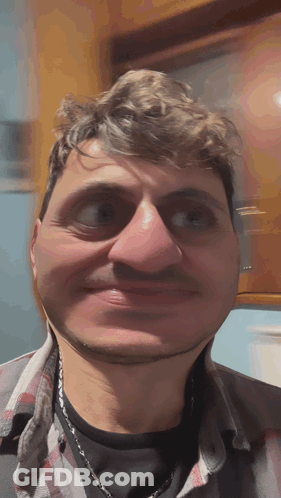 A Man Wearing A Plaid Shirt And A Necklace Makes A Funny Face GIF