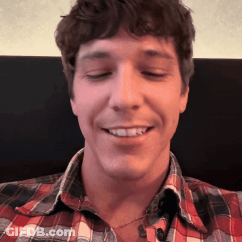A Man Wearing A Plaid Shirt And A Necklace Smiles With His Eyes Closed GIF