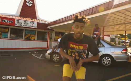 A Man Wearing A Pulp Fiction T-Shirt Stands In Front Of A Mike 'S Restaurant GIF