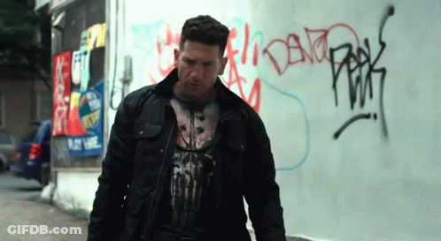 A Man Wearing A Punisher T-Shirt Stands In Front Of A Graffiti Covered Wall GIF