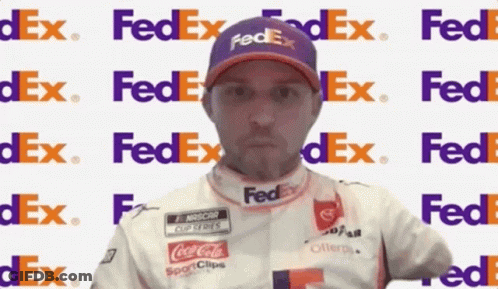 A Man Wearing A Purple Fedex Hat Stands In Front Of A Fedex Logo GIF