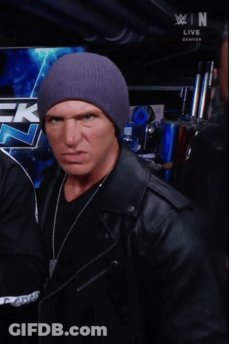 A Man Wearing A Purple Hat Is Making A Funny Face In Front Of A Wrestling Logo GIF