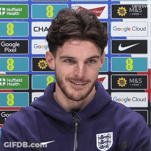 A Man Wearing A Purple Hoodie Stands In Front Of A Wall Of Advertisements Including Google Pixel And M & S Food GIF