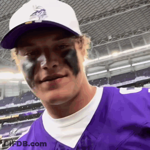 A Man Wearing A Purple Vikings Jersey And A White Hat Has Black Paint On His Face GIF